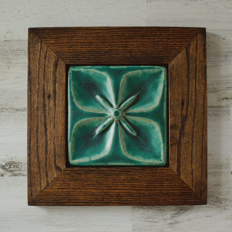 The Flower Geo Tile features a four-petaled flower. Each petal takes up a quarter of the tile, making a perfectly geometric shape. The inner part of the flower is round with four cattail shaped stamen exploding from the center that point out to the corners of the tile. This tile is framed in a cool colored wood.