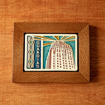 The Guardian Building Tile design features the top of the building on the right side of the tile with the word "Guardian" on the left. A large version of the Pewabic tiles found on the Guardian Building are framing the left side of the image. The tile is hand-painted in blues with red and yellow accents. The oak wood frame is stained with a yellowy brown finish.
