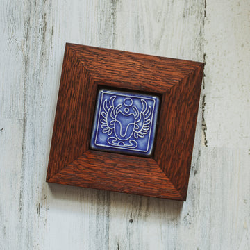 The Scarab Tile features a line drawing of a scarab beetle with wings outstretched and two small circles floating between its front arms. The design has classic Egyptian markings. This tile is in the glossy bright blue Lake Superior glaze which beautifully offsets the deep reddish brown oak wood frame.