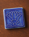A glossy blue tile with an embossed line drawing of a lotus flower.