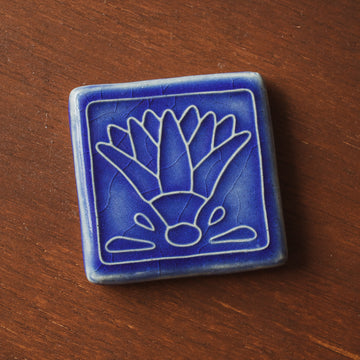 A glossy blue tile with an embossed line drawing of a lotus flower.