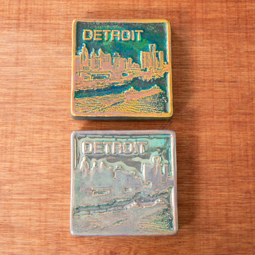Two metallic iridescent tiles are shown with vastly different finishes. The Detroit tile design includes the Detroit skyline as seen from the Detroit river. There is a freighter skating over the water, causing ripples. The word DETROIT floats above the skyscrapers.
