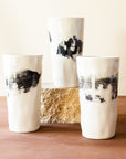 Three tall Kirsten Helmer cups stand together, each is glossy white with thick black paint swiped around its waist.