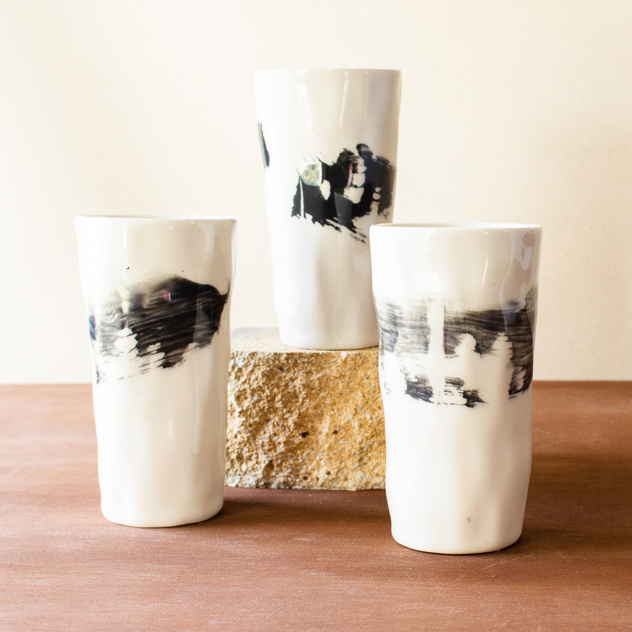 Three tall Kirsten Helmer cups stand together, each is glossy white with thick black paint swiped around its waist.