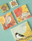 Five vibrantly colored tiles featuring line drawings of birds and flowers lay together on a flat, painted surface.
