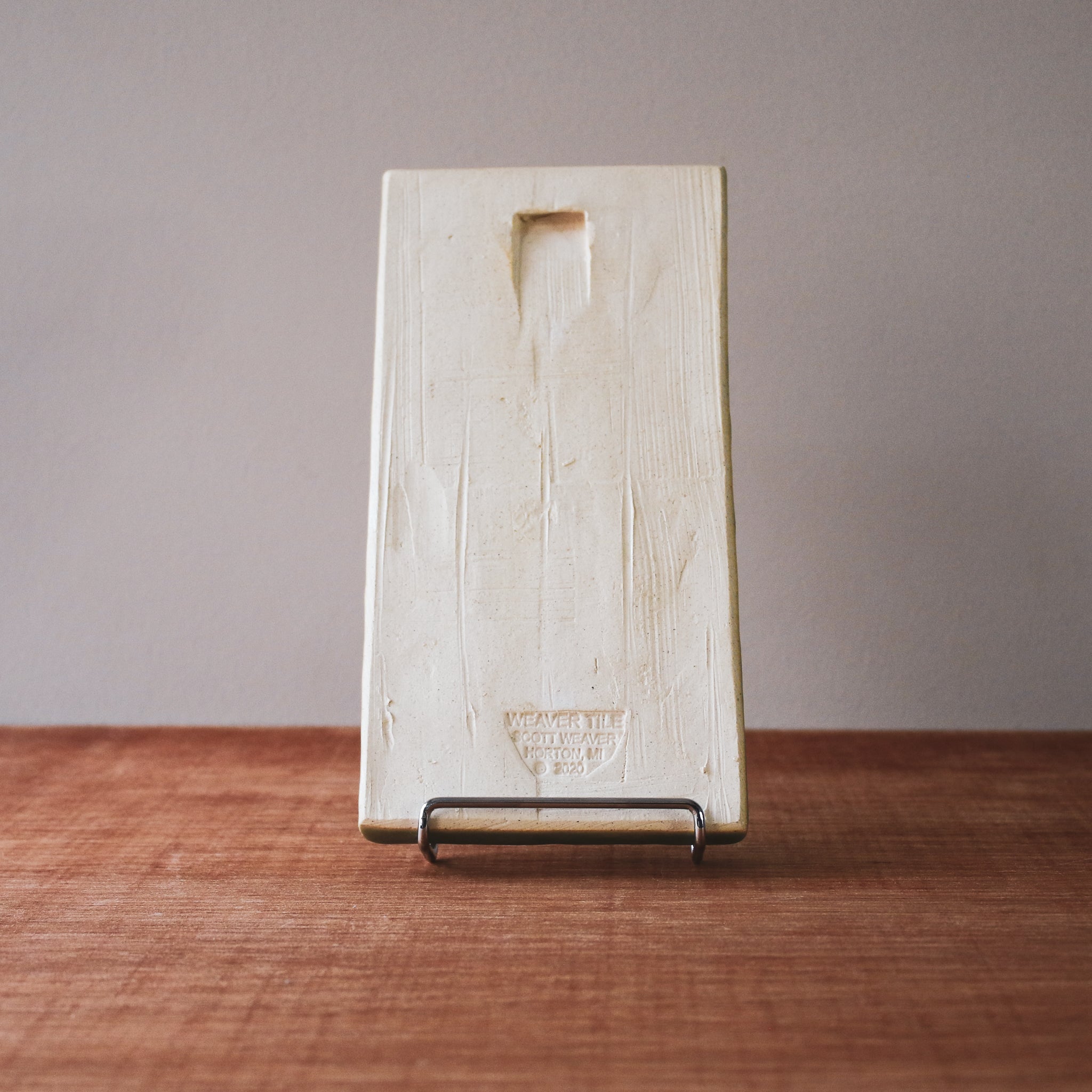 The back of the tile has a divot in the clay making is possible to hang it from a nail. There is also a stamp from the maker saying "Weaver Tile Scott Weaver Horton, MI".