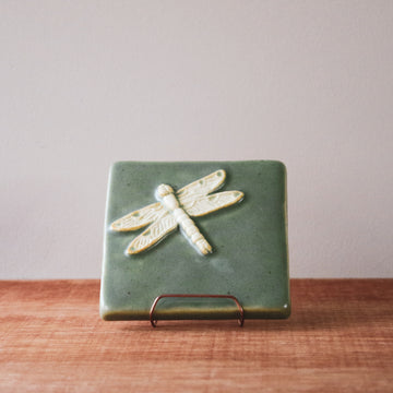 This high relief tile features a dragonfly with detailed wings. It seems to be flying across the tile and sits diagonally, looking to the top right of the tile. The dragonfly is scraped white while the background is a light green color.