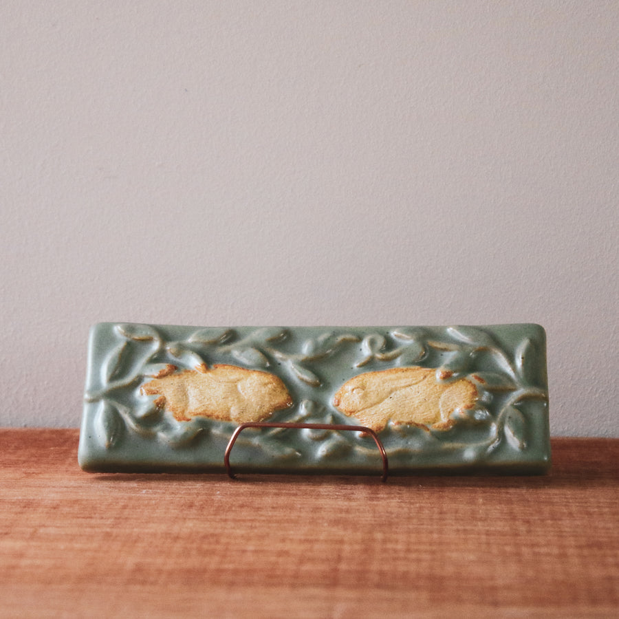 This high relief tile features two young rabbits, resting with ears flat against their backs. They are nestled amongst the green leaves of a protective bush. The rabbits are in scraped of glaze giving them a creamy white color while the rest of the tile is a pale green.