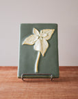 This high relief rectangular tile features a large trillium flower. The three petalled flower has a trio of large leaves under it and a slightly curved stem. The flower is scraped to make a creamy white color while the background is a pale green.