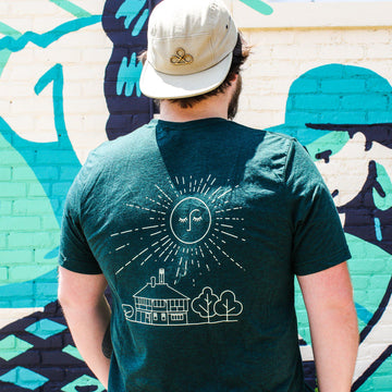 The back of this deep green t-shirt has a large yellow illustration of the Pewabic building surrounded by bushy trees. Above them a large sun with closed eyes and a nose is spreading its rays outward.