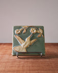 This tile features a thin branch with two flowers blooming on it. A hummingbird with wings outstretched reaches its beak to wards the blooms. The bid and flowers are scraped creamy white and the background is a pale green color.