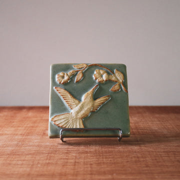This tile features a thin branch with two flowers blooming on it. A hummingbird with wings outstretched reaches its beak to wards the blooms. The bid and flowers are scraped creamy white and the background is a pale green color.