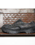 This multimedia sculpture features a life-like ceramic shoe. It is black and worn with missing shoelaces and a thick grippy sole. The work shoe sits on a corrugated metal shelf with decorative gears that like silver metal that is rusting.