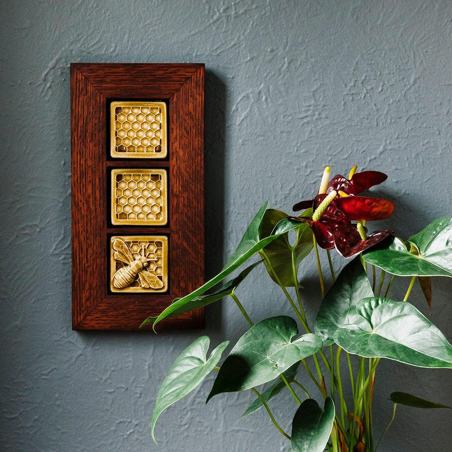 This framed triptych features one Honey Bee Tile and two Honeycomb tiles. The Honeybee Tile features a large honeybee with stripes and detailed segmented wings sitting on a honeycomb patterned background. The Honeycomb tiles are a flat line drawing of a honeycomb pattern with a simple smooth border around it. These three tiles are lined up and attached in a deep reddish brown oak frame.