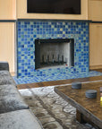 A slightly angled photograph of a blue iridescent tiled fireplace. The tile looks very vibrant from this perspective. There is a gray, velvet-looking couch and dark wood coffee table with gray coasters in view.