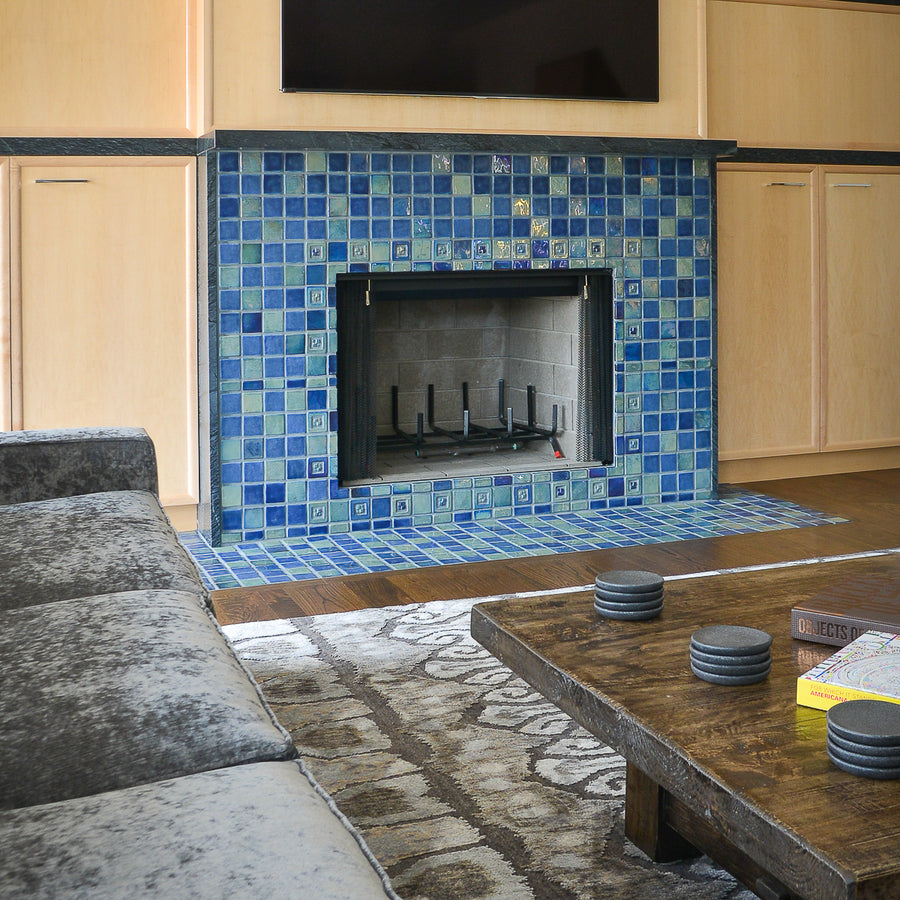 A slightly angled photograph of a blue iridescent tiled fireplace. The tile looks very vibrant from this perspective. There is a gray, velvet-looking couch and dark wood coffee table with gray coasters in view.