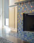 Angled photograph of a blue iridescent tiled fireplace. There is a matte charcoal vase in view. The fireplace is surrounded by light wood cabinets. You can see a small corner of an ornate, gray rug on the floor in front of the fireplace. 