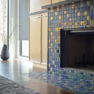 Angled photograph of a blue iridescent tiled fireplace. There is a matte charcoal vase in view. The fireplace is surrounded by light wood cabinets. You can see a small corner of an ornate, gray rug on the floor in front of the fireplace. 