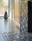 Angled photograph of a blue iridescent tiled fireplace. There is a matte charcoal vase in view. The fireplace is surrounded by light wood cabinets. 