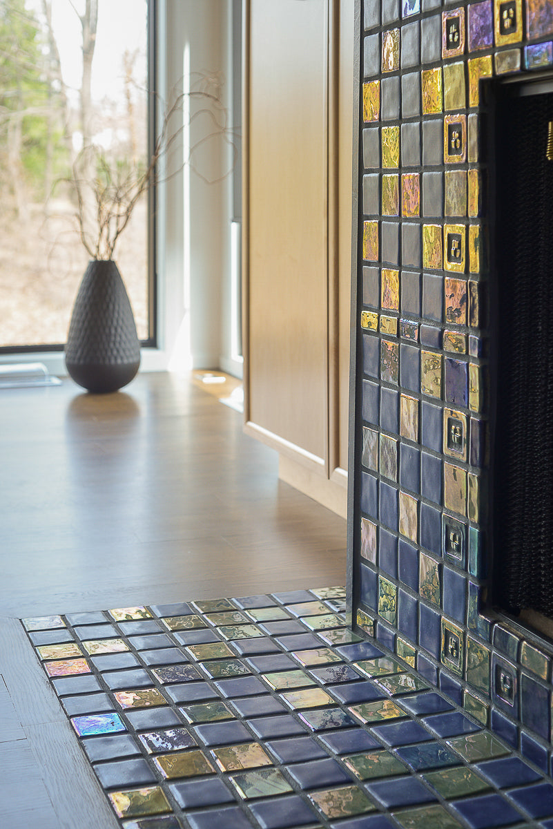 Angled photograph of a blue iridescent tiled fireplace. There is a matte charcoal vase in view. The fireplace is surrounded by light wood cabinets. 