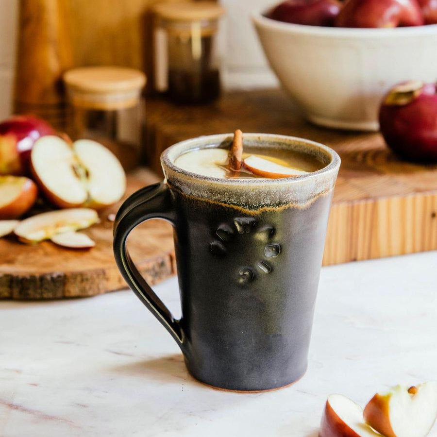 Pewabic Cafe Mug in our matte-black Carbon glaze. The mug is filled with mulled apple cider on a marble countertop. There is a Pewabic bowl in a white Birch glaze filled with red apples in the background.