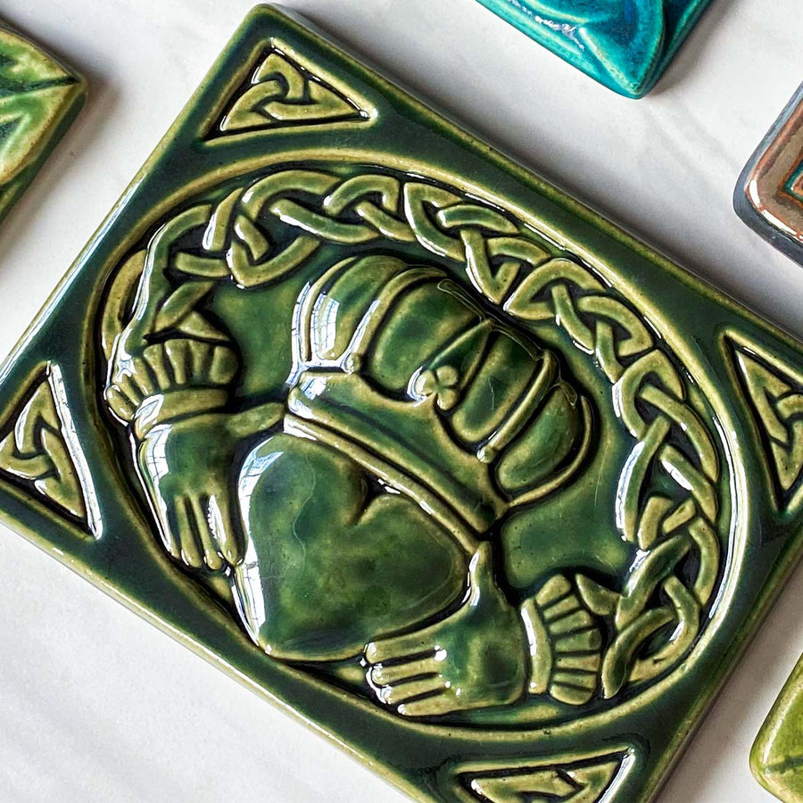 This Claddagh tile features the standard heart, hands and crown design in the center with a Celtic knot motif that wraps above the design and connects the hands- harkening to the original purpose of the Claddagh, as a ring. This tile features the glossy deep green Kale glaze.