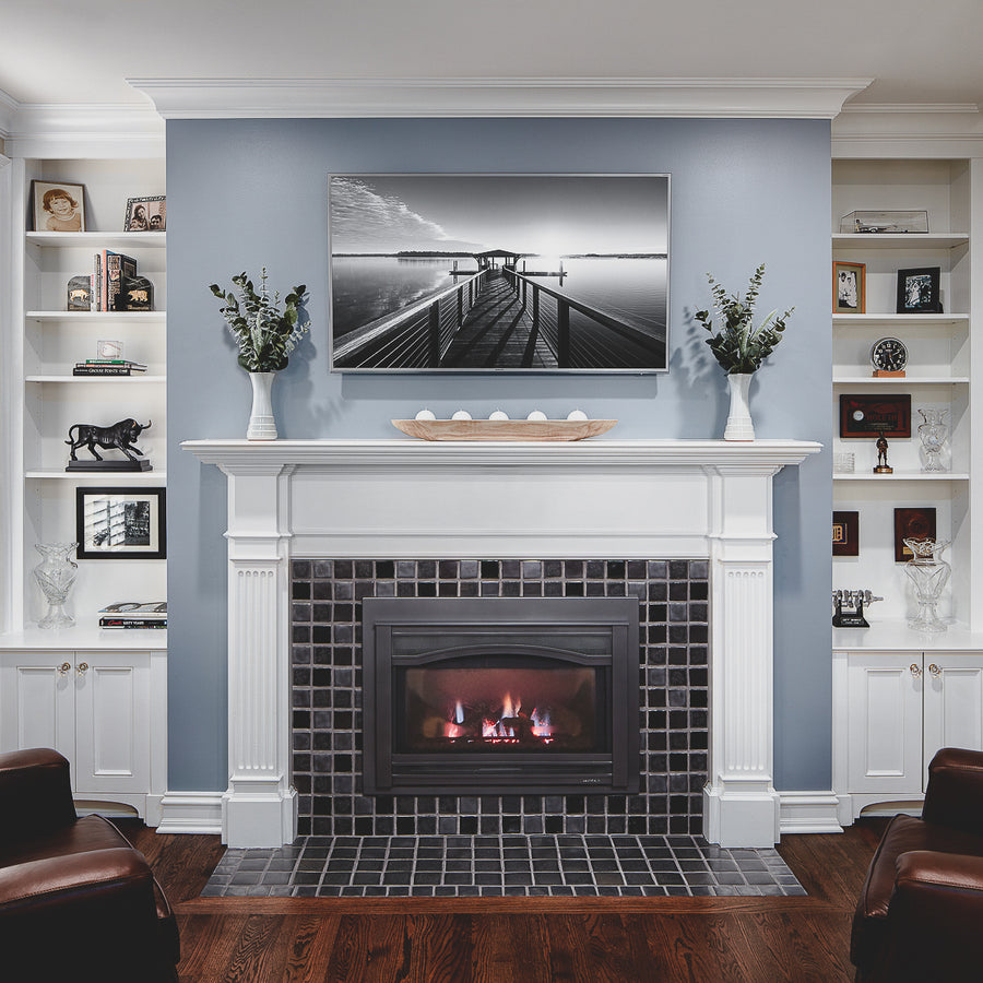 A straight on view of a fireplace and hearth made up of charcoal and glossy black or "onyx" tiles. There is a black and white photo of a dock above the white mantel which holds two "Birch" glazed Pewabic Step Vases. The surrounding white shelves contain personal items such as photos and statues. There are two dark-brown leather couches coming into the corner of the photo. 
