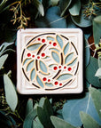 The DIA Leaf and Berries tile features two intertwined branches with round berries that create a circle at the center of the tile. The thick lined border makes the tile into a square. This tile features Style D with red berries, pale green leaves, brown stems and a white background.