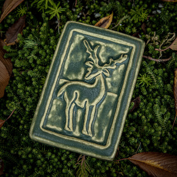 Our 4" x 6" Deer Tile in a muted green "Bayleaf" glaze. You can see the outline of the deer with antlers, which is contrasted a pale-yellow tone. It is laying on a bed of small evergreens surrounded by crisp, brown, autumn leaves.