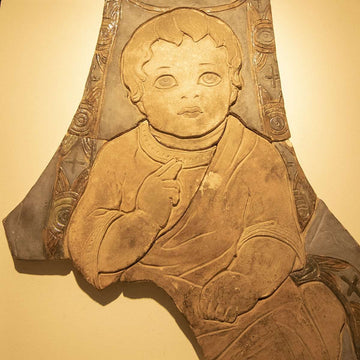 This partial tile installation features the baby Jesus wearing robes with one chubby arm resting on his lap while the other has his pointer finger and middle finger up and resting on his chest. His eyes are pointed upward.