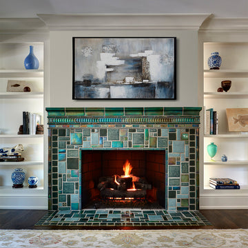 Vibrant, lit fireplace made up of Iridescent and matte glazes which creates an overall green, mirror-like effect. The white shelves on either side of the fireplace hold various ceramic pieces–– mostly in bright blue and ivory. There is a blue, gray, and white abstract painting above the mantel.