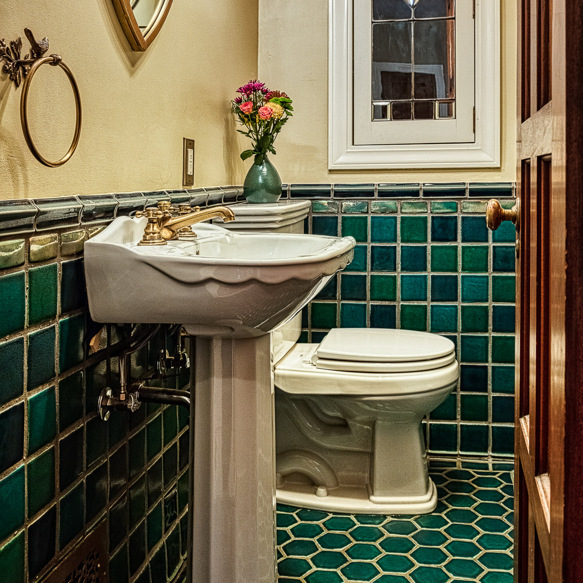A wooden door opens to a custom tiled bathroom in varying greens and blues. The walls are an off-white color and the bathroom accents colors are mostly white and brass. 