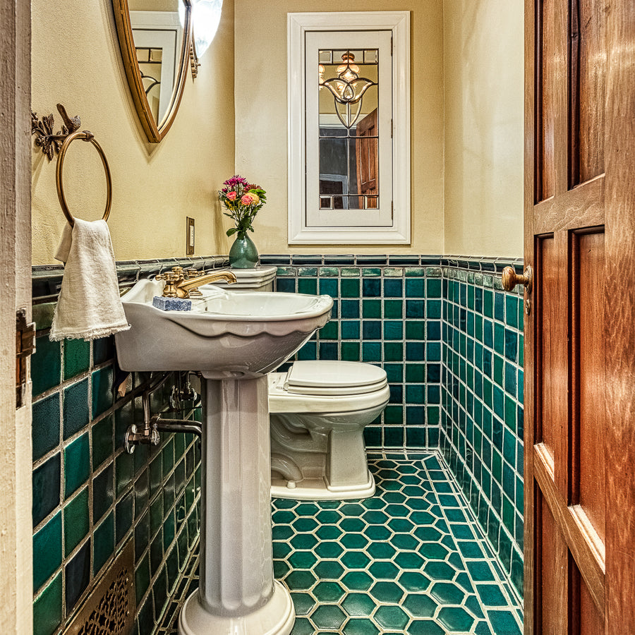 A wooden door opens to a custom tiled bathroom in varying greens and blues. The walls are an off-white color and the bathroom accents colors are mostly white and brass. 