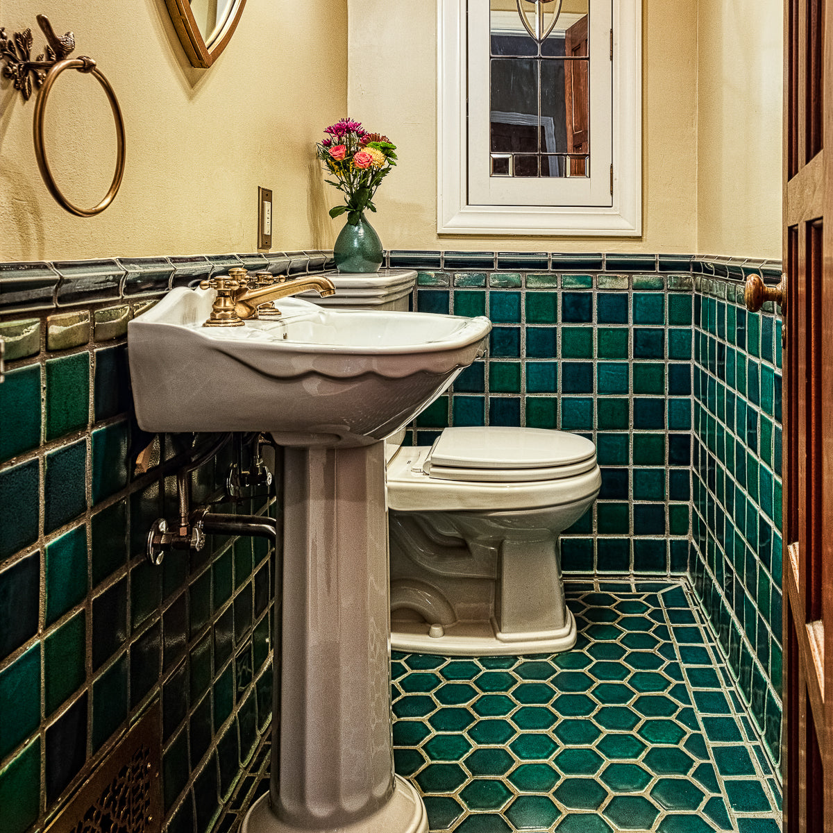 A wooden door opens to a custom tiled bathroom in varying greens and blues. The walls are an off-white color and the bathroom accents colors are mostly white and brass. 