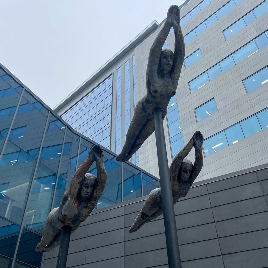 Three Swimmer sculptures are raised above the ground on metal poles. Each swimmer is a nude woman - their bodies are flat and straight with their stomachs facing down and their arms and legs pointed as if diving through the water.