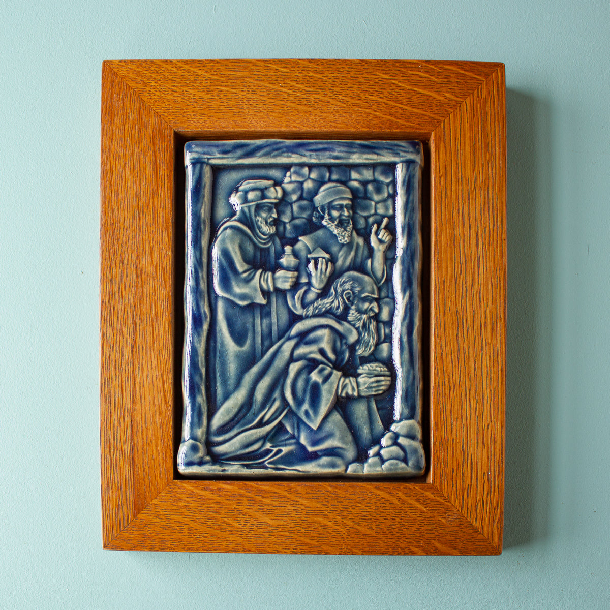 The Three Wise Men Tile features the wisemen in robes holding their three gifts- gold, frankincense and myrrh. They are all facing the right, they seem to be focused on one point. There is a stone wall behind them. This tile is in the glossy deep blue Ocean glaze which offsets the pale yellowish brown oak wood frame.