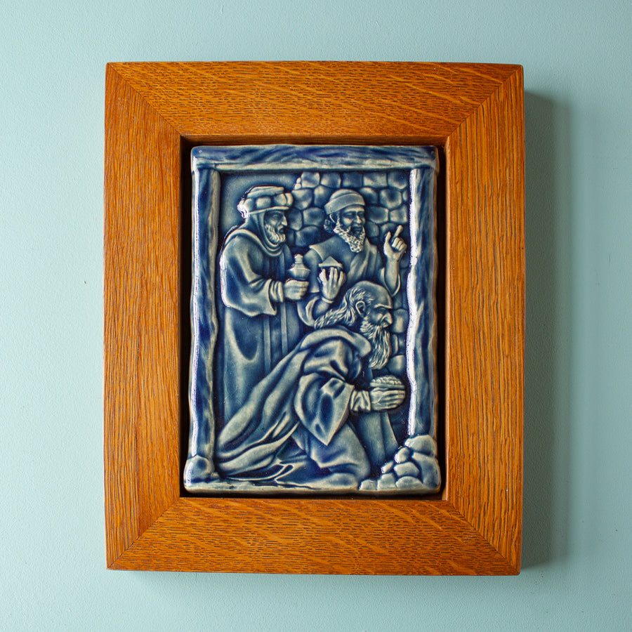 The Three Wise Men Tile features the wisemen in robes holding their three gifts- gold, frankincense and myrrh. They are all facing the right, they seem to be focused on one point. There is a stone wall behind them. This tile is in the glossy deep blue Ocean glaze which offsets the pale yellowish brown oak wood frame.