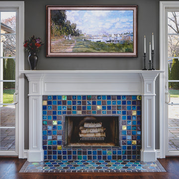 This fireplace features Iridescent tile accents along with a mix of complementary blue and charcoal tones. There are three black candlesticks and a charcoal glazed Pewabic vase on the Colonial style, white mantel. Above the mantel there is a painting of a seaside town.  