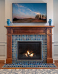 A lively mix of blue, gray, and charcoal tiles are used in this fireplace and hearth set in a Craftsman style wooden mantel. There are two Pewabic Classic Vases on either side of the mantel in our true blue "Peacock" glaze. The fire is lit and there is a landscape photo framed on the wall of a sand dune with wispy trees in the background. 