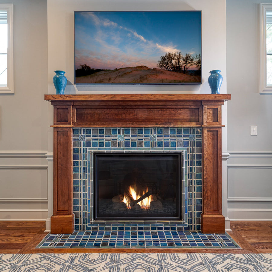 A lively mix of blue, gray, and charcoal tiles are used in this fireplace and hearth set in a Craftsman style wooden mantel. There are two Pewabic Classic Vases on either side of the mantel in our true blue "Peacock" glaze. The fire is lit and there is a landscape photo framed on the wall of a sand dune with wispy trees in the background. 