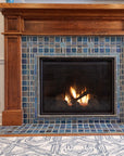 A lively mix of blue, gray, and charcoal tiles are used in this fireplace and hearth set in a Craftsman style wooden mantel. There are two Pewabic Classic Vases on either side of the mantel in our true blue "Peacock" glaze. The fire is lit and there is a gray and white geometric rug in view on the floor.