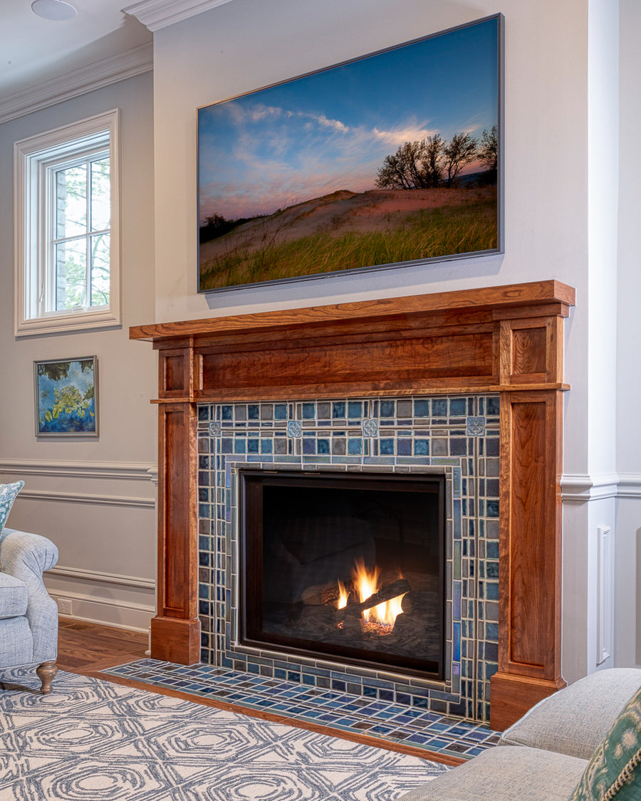 A lively mix of blue, gray, and charcoal tiles are used in this fireplace and hearth set in a Craftsman style wooden mantel.  The fire is lit and there is a landscape photo framed on the wall of a sand dune with wispy trees in the background.