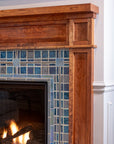 A detail shot of a fireplace made up of blue, gray, and charcoal tiles. The camera is focused on a corner of the fireplace, which shows the wood grain detail of the Craftsman style mantel surround.