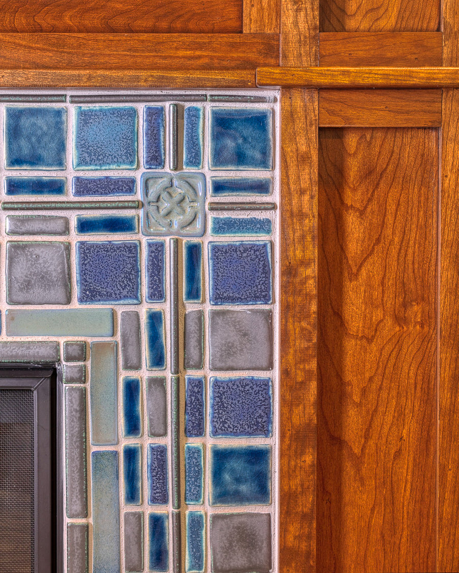 A detail shot of a fireplace and hearth made up of blue, gray, and charcoal tiles. The camera is focused on a corner of the fireplace, which shows the wood grain detail of the Craftsman style mantel surround.