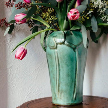 The Lotus Vase features a tall base with long stalks reaching up to a ring of lily pads that crown the top and lip of the vase.