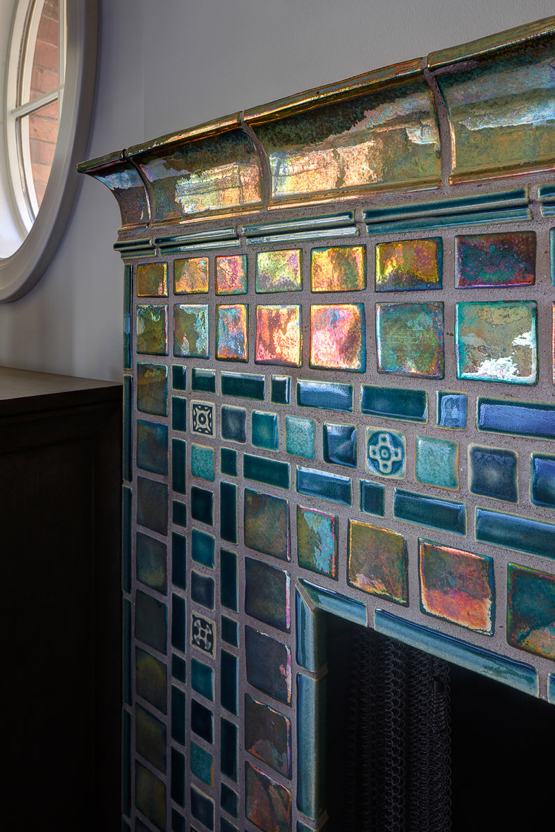 A corner detail shot of a neo-futuristic fireplace and hearth comprised of Iridescent tiles which are green/blue and mirror-like. 