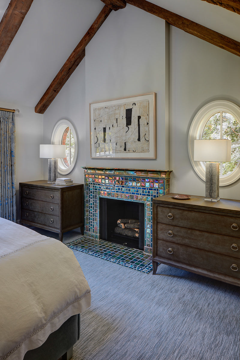 A neo-futuristic fireplace and hearth comprised of Iridescent tiles which are green/blue and mirror-like. There is a framed piece of abstract art above the fireplace with wooden dressers, round windows, and lamps on either side.