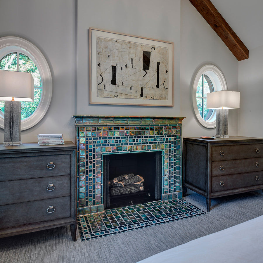 A neo-futuristic fireplace and hearth comprised of Iridescent tiles which are green/blue and mirror-like. There is a framed piece of abstract art above the fireplace with wooden dressers, round windows, and lamps on either side.