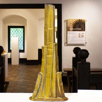 This tall sculpture is shaped like a skinny volcano with straight sides that get wider as steps of gilded clay are added to its surface. It is colored in yellows and browninsh grays.