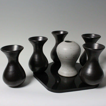 A small black platter holds five shiny black vases- they are smooth and curved. Nestled between them is a white vase with a speckled surface.